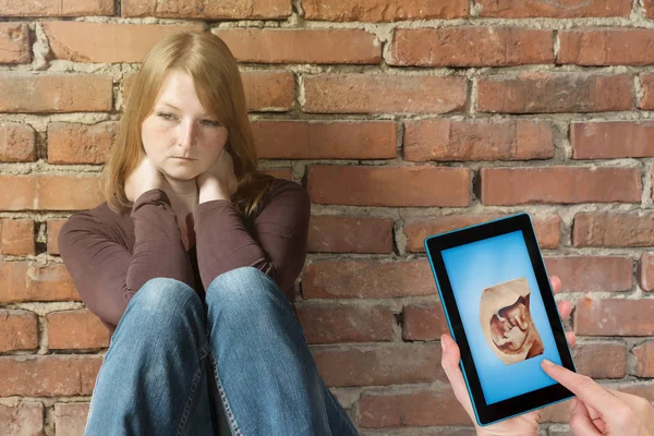 悲伤的年轻女子坐在旧砖墙前 另一只女性手在平板电脑屏幕上展示未出生孩子的空间镜头 — 图库照片