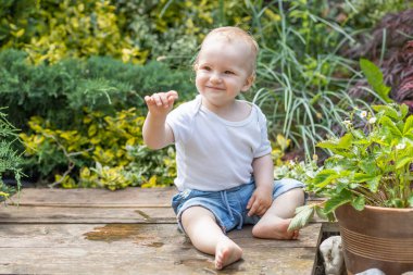 Gülümseyen küçük oğlan bahçesinde bitkiler olan bir saksının yanında oturuyor.