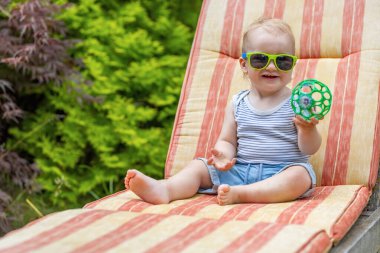 Güneş gözlüğü takan gülümseyen küçük çocuk güvertedeki sandalyede oturan oyuncak topu gösteriyor..
