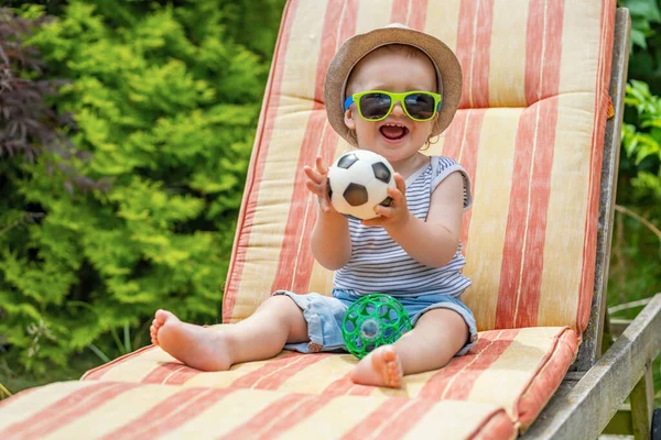 Güneş gözlüğü takan gülen küçük çocuk bahçede iskemlede oturan toplarla oynuyor..