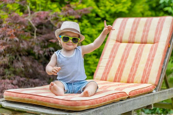 Güneş gözlüğü takan gülümseyen bebek bahçede iskemlede işaret parmağıyla oturuyor..