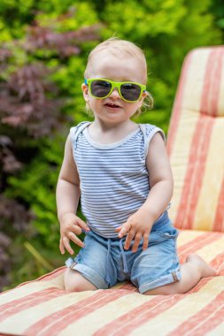 Güneş gözlüğü takan küçük bebek bahçede iskemlede diz çöküyor. Dikey.
