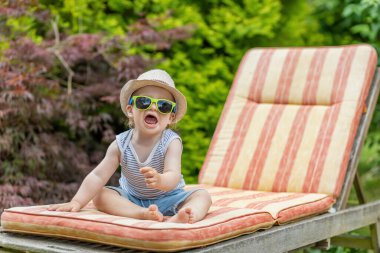 Güneş gözlüğü takan küçük bebek bahçede iskemlede kameraya bağırıyor..