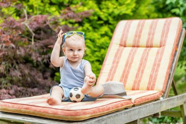 Küçük bebek terasta, elinde güneş gözlüğü ve bacağıyla poz veriyor..