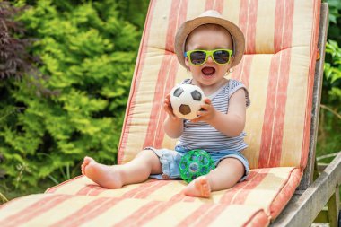 Ağzı açık, güneş gözlüğü takan küçük mutlu bebek elinde küçük bir futbol topu oyuncağıyla güvertede oturuyor..