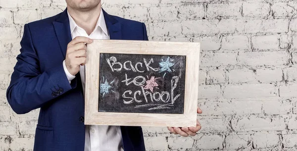 Man in classic suit holds blackboard with title Back to school over white bricks background. No face, horizontal banner, copy space. Start of school year concept.