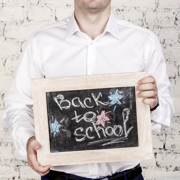 Man in white shirt holds blackboard with title Back to school over white bricks background. No face, Start of school year concept, square picture.