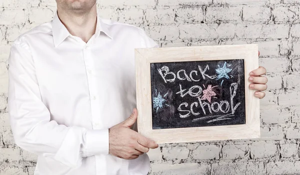 Man in white shirt holds blackboard with title Back to school over white bricks background. No face, Start of school year concept, horizontal banner.