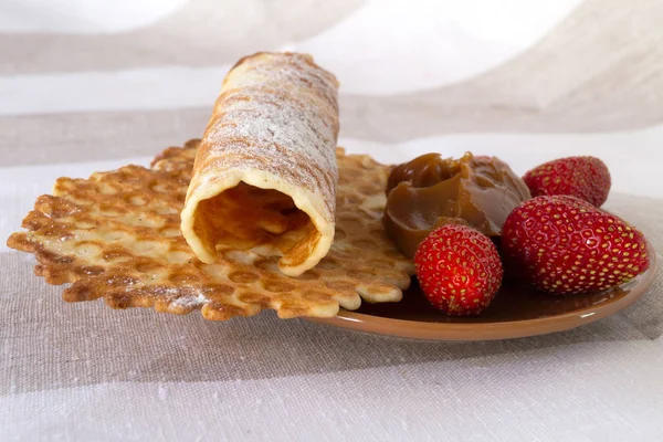 Round waffles and a roll of thin short pastry with chocolate pasta and strawberries on a round plate.