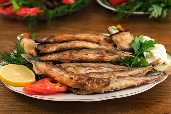 Small fish capelin, fried in oil until golden  brown, laid on a plate with rings of sweet pepper, a slice of lemon, parsley and dill.