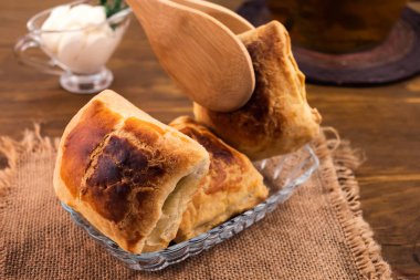 Bir cam levha Mutfak maşa, kaba keten kumaş, et suyu tekne ile beyaz ekşi krema, ahşap arka plan üzerinde bir yakın çekim yapılmış bir peçete ile yerleştirildiği puf çörekler kırmızı bir kabuk ile.