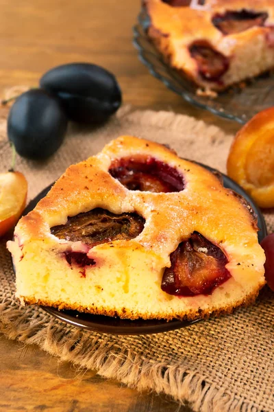 Cut off a piece of homemade plum cake, stacked on a round plate, ripe whole plums on a linen napkin, vertical frame.