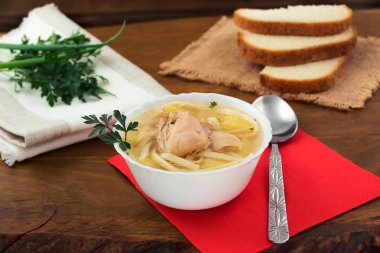 Beyaz bir kase, bir çorba kaşığı kırmızı peçeteye, beyaz ekmek, dilim tavuk şehriye bir kısmı taze yeşil maydanoz.