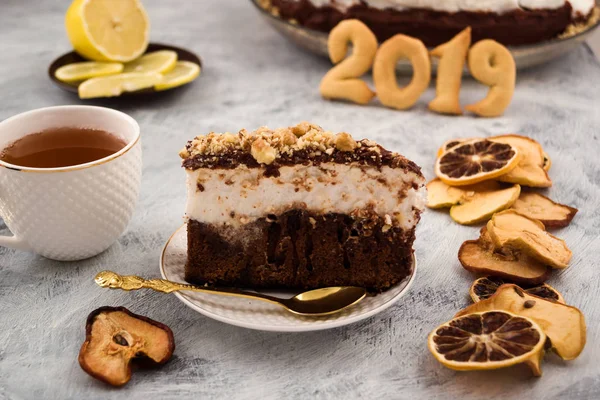 Festive dessert. Piece of chocolate cake. White meringue, brown dough. Figures 2019. Dry fruit slices. White mug with a drink. Sliced fresh lemon. Light background.