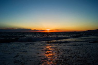 Akşam güneşi günbatımı sırasında denizde. Küçük dalgalar ve köpük ile plaj. Güneş ışığı su yansıması.
