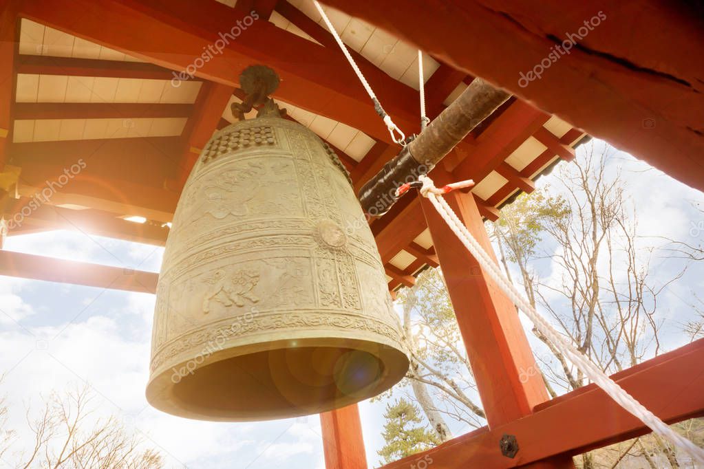 Big brass Buddhist bell and timber knock of Japanese temple in red pavilion on bright blue sky with sun and lens flare background.