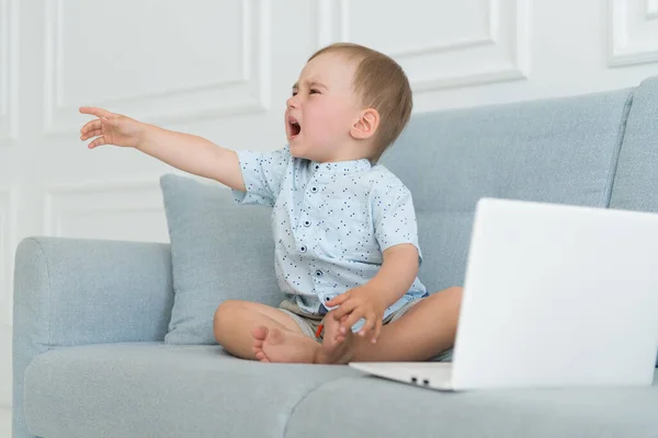 悲しい赤ちゃんがノートパソコンの近くで泣いています ソファの上のタンタル子 — ストック写真