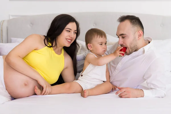 happy family on bed in the bedroom married couple with small child and pregnant woman - baby feeds father