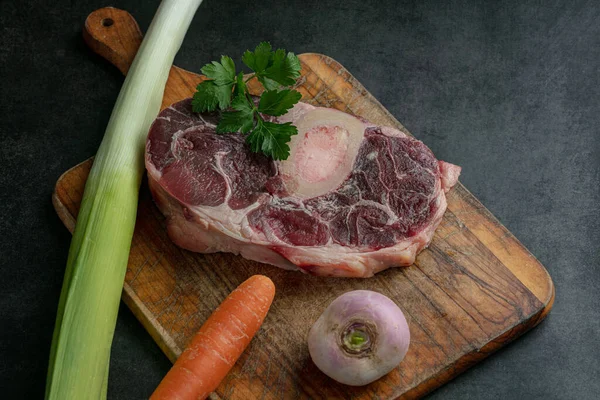 Slice of raw beef shank with vegetables on a cutting board on a dark background
