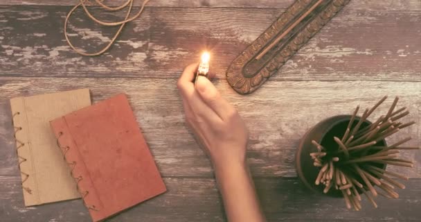 La mano de mujer enciende una varilla de incienso de un sostenedor de buda en una superficie de madera natural vintage con libros, cordel de cáñamo y muchas varillas de incienso aromáticas. Concepto de ritual espiritual, ideas de creatividad o imaginación. — Vídeo de stock