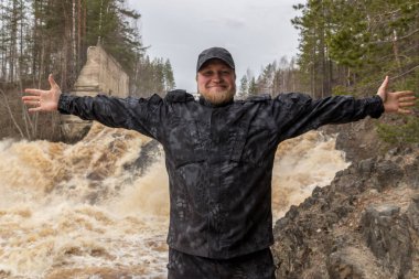 Fırtınalı bir nehrin arka planına karşı siyah üniformalı göğsüne katlanmış kolları olan bir sakallı adam