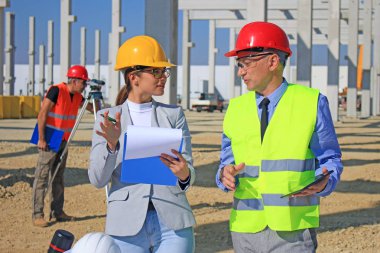 İnşaat alanındaki projeden bahseden mimar ve inşaat mühendisi kadın. Arkalarında ise ölçüm cihazı ve takım çalışması olan inşaat işçisi var.