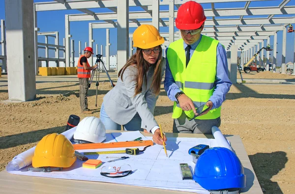 建設現場でプロジェクトについて話すハードハットの女性建築家と建設エンジニア 測定装置 チームワークで建設労働者の背後にある — ストック写真