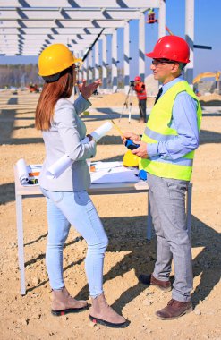 İnşaat alanındaki projeden bahseden mimar ve inşaat mühendisi kadın, arkalarında ölçüm cihazı, vinç ve diğer inşaat işçileriyle birlikte inşaat işçisi, takım çalışması