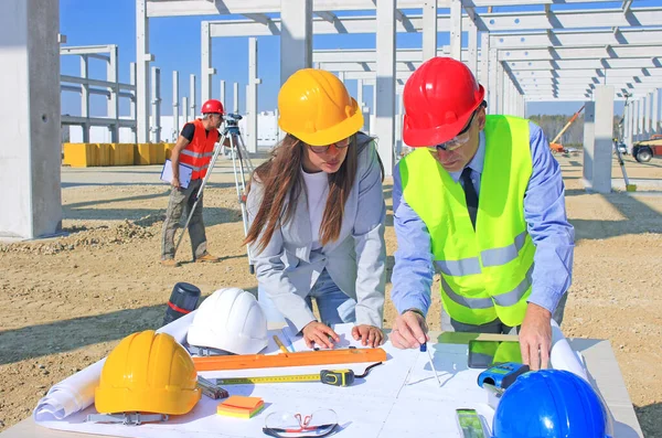 戴着硬礼帽的女建筑师和建筑工程师在建筑工地谈论这个项目 后面是带着测量装置和团队合作的建筑工人 — 图库照片