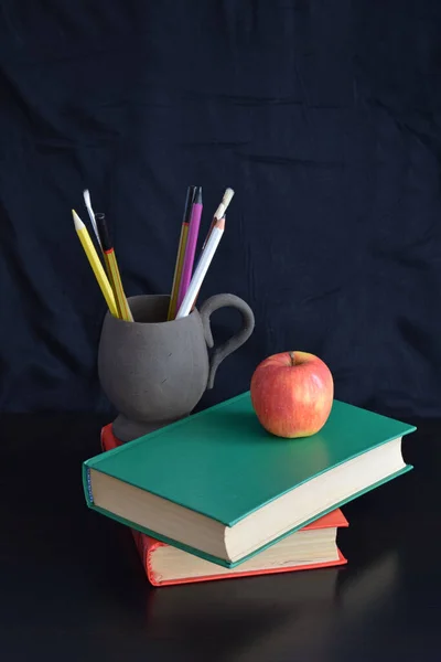 Books and an Apple on a black background.Pencils in a clay Cup.