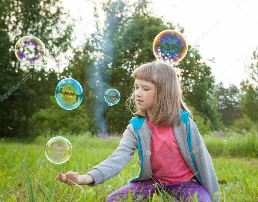 Cute preschooler girl playing with soap bubbles sitting in a summer park