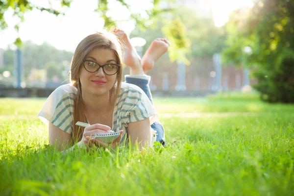 美丽的年轻女子在日记中做笔记躺在一个阳光明媚的草地上 — 图库照片