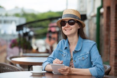 Bir fincan kahve ile kafede oturan ve günlükte notlar alarak gülümseyen genç kadın