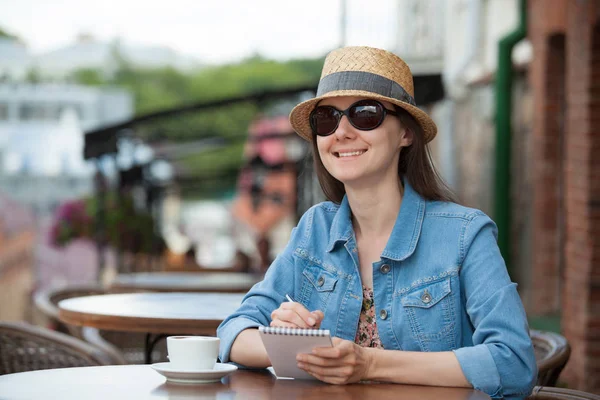 Bir fincan kahve ile kafede oturan ve günlükte notlar alarak gülümseyen genç kadın