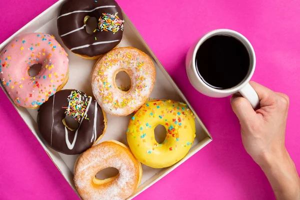Donuts in a box on a pink background.Hand holding cup of coffee. Top of view.