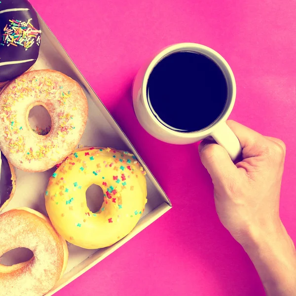 Donuts in a box on a pink background.Hand holding cup of coffee. Toned, Top of view.