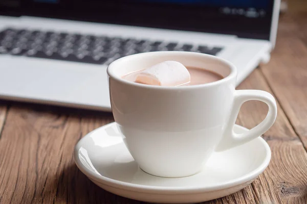 Cup of cocoa with marshmallow and laptop on a wooden background