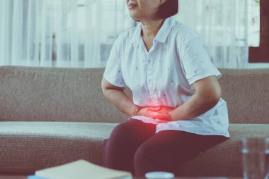 Yaşlı Asyalı bir kadın evdeki koltukta otururken midesi ağrıyor. Yaşlı bir kadın karın ağrısı çekiyor.