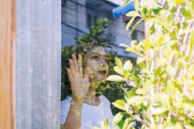 Asyalı kadın elleri cama dokunur, Coronavirüs ve covid-19 salgını sırasında evde kalır veya salgın hastalık, sosyal uzaklık kavramı
