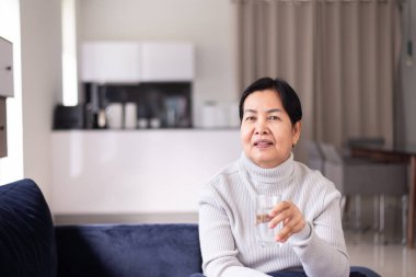 Kıdemli Asyalı kadın sabah evde su içiyor, mutlu ve gülümsüyor, yaşlı sağlıklı bakım konsepti.