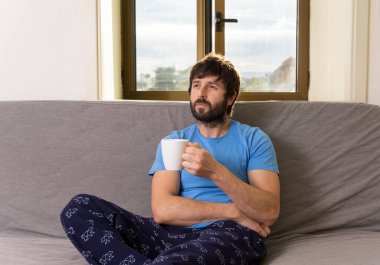 Oturma odasındaki kanepede elinde fincanla oturan genç, havalı bir adam. Düşünceli ifade