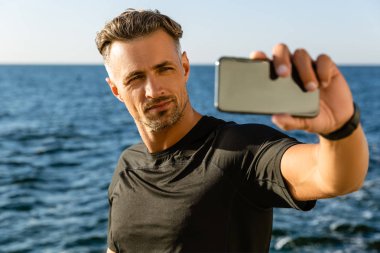 handsome adult man taking selfie with smartphone on seashore clipart
