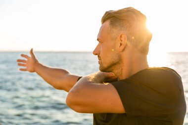 handsome adult sportsman stretching arm before training on seashore in front of sunrise clipart
