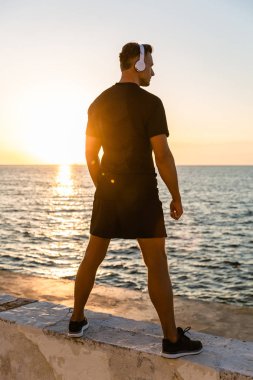 rear view of athletic adult man in headphones standing on seashore in front of sunrise clipart