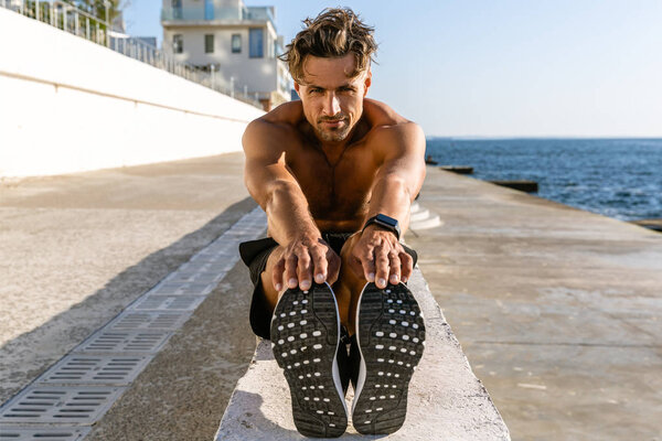 muscular adult shirtless sportsman stretching before training on seashore