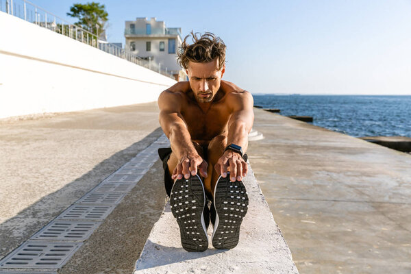 adult shirtless sportsman stretching before training on seashore