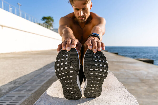 close-up shot of adult shirtless sportsman stretching before training on seashore