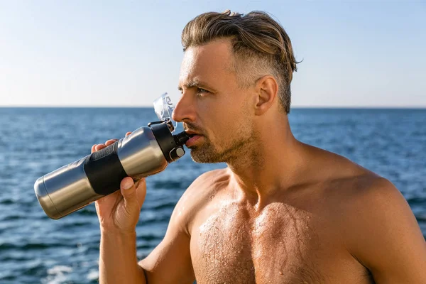 shirtless handsome adult man drinking water after workout on seashore