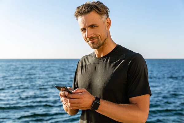 handsome adult man with smartphone on seashore and looking at camera
