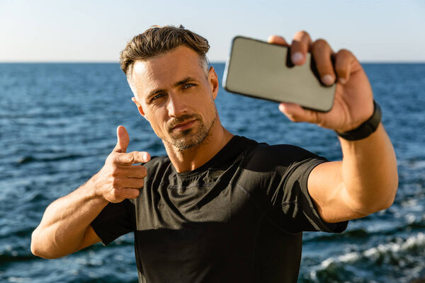 handsome adult man taking selfie with smartphone and pointing at camera on seashore